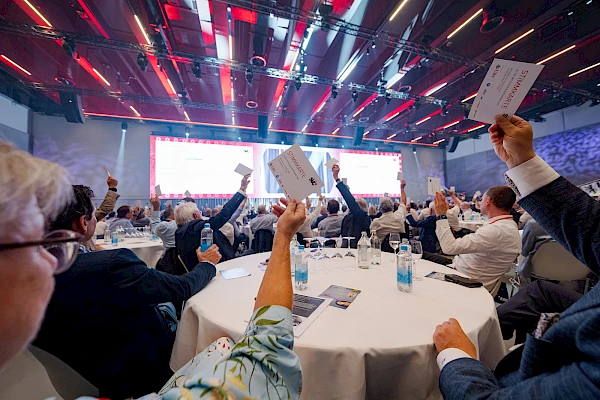 Eventfotograf Festhalle Bern: Abstimmung an einer Generalversammlung – Gäste an runden Tischen heben ihre Stimmkarten hoch, im Hintergrund die Präsentationsleinwand.